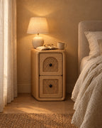 Narrow natural wood bedside table with two woven cane drawers, a lamp switched on and an open book in the evening