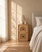 Narrow natural wood bedside table with two woven cane drawers with a ceramic lamp in morning light