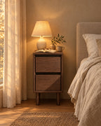 Narrow walnut-effect bedside table with a lamp switched on and an open book in the evening