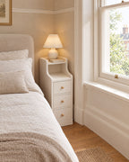 Narrow 20 cm white bedside table beside a linen bed in a British bedroom with a sash window