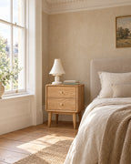 Oak two-drawer bedside table on tapered legs beside a linen bed in a British bedroom with a sash window