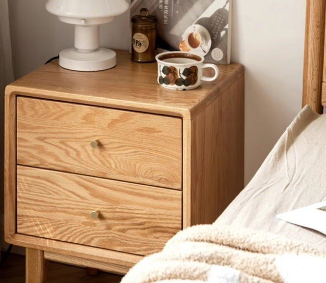 Oak bedside table with two drawers and brass knobs beside a bed