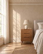Oak bedside table with three drawers beside a linen bed in a bright British bedroom with a sash window