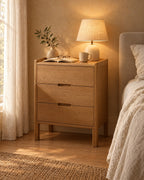 Oak bedside table with three drawers, a lamp switched on and an open book in the evening
