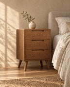 Oak bedside table with three drawers standing beside a bed with linen bedding