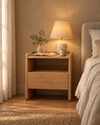 Oak bedside table with an open shelf and one drawer, a lamp switched on and an open book in the evening