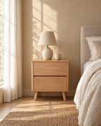 Oak bedside table with two drawers and tapered legs with a ceramic lamp in morning light