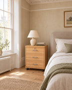 Oak-effect three-drawer bedside cabinet on castors beside a linen bed in a British bedroom with a sash window