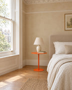 Orange metal pedestal side table beside a linen bed in a bright British bedroom with a sash window