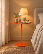 Orange metal pedestal side table with a lamp switched on and an open book in the evening