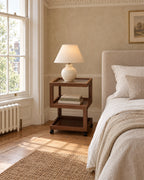 Walnut-effect rolling bedside table with three open tiers beside a linen bed in a bright British bedroom with a sash window