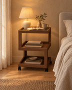 Walnut-effect rolling bedside table with three open tiers with a lamp switched on and an open book in the evening