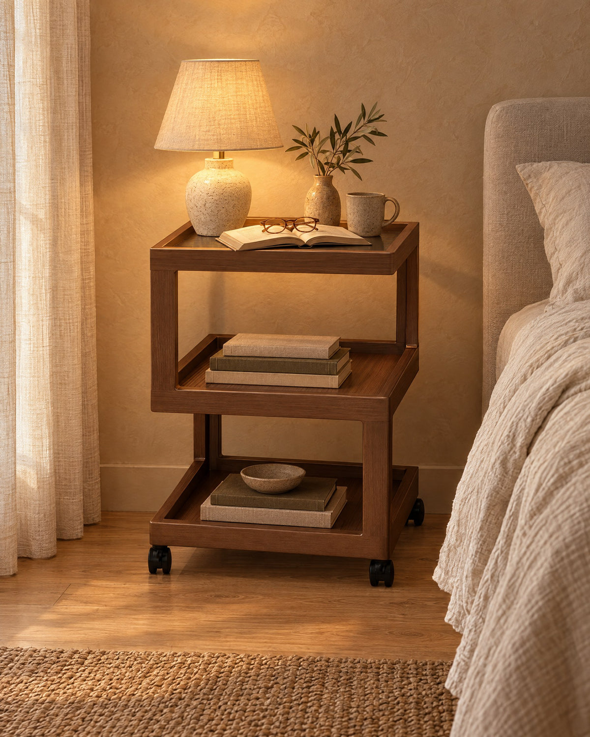 Walnut-effect rolling bedside table with three open tiers with a lamp switched on and an open book in the evening