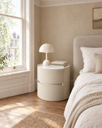 Round cream bedside table beside a linen bed in a British bedroom with a sash window