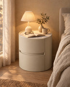Round cream bedside table with a stone top and a lamp switched on in the evening