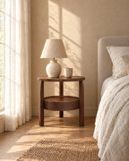 Round walnut-tone bedside table with three legs with a ceramic lamp in warm morning light
