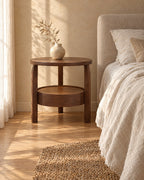 Three-quarter view of the round walnut-tone bedside table with three legs beside a bed