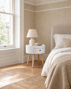 Round white bedside table with natural wood legs beside a linen bed in a bright British bedroom with a sash window