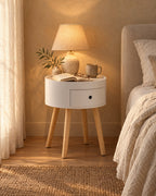 Round white bedside table with natural wood legs with a lamp switched on and an open book in the evening