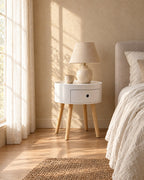 Round white bedside table with natural wood legs in warm morning light with window shadows on the wall