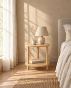 Round two-tier wooden bedside table with a ceramic lamp in morning light