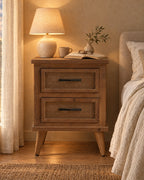 Rustic wood bedside table with herringbone drawers with a lamp switched on and an open book in the evening