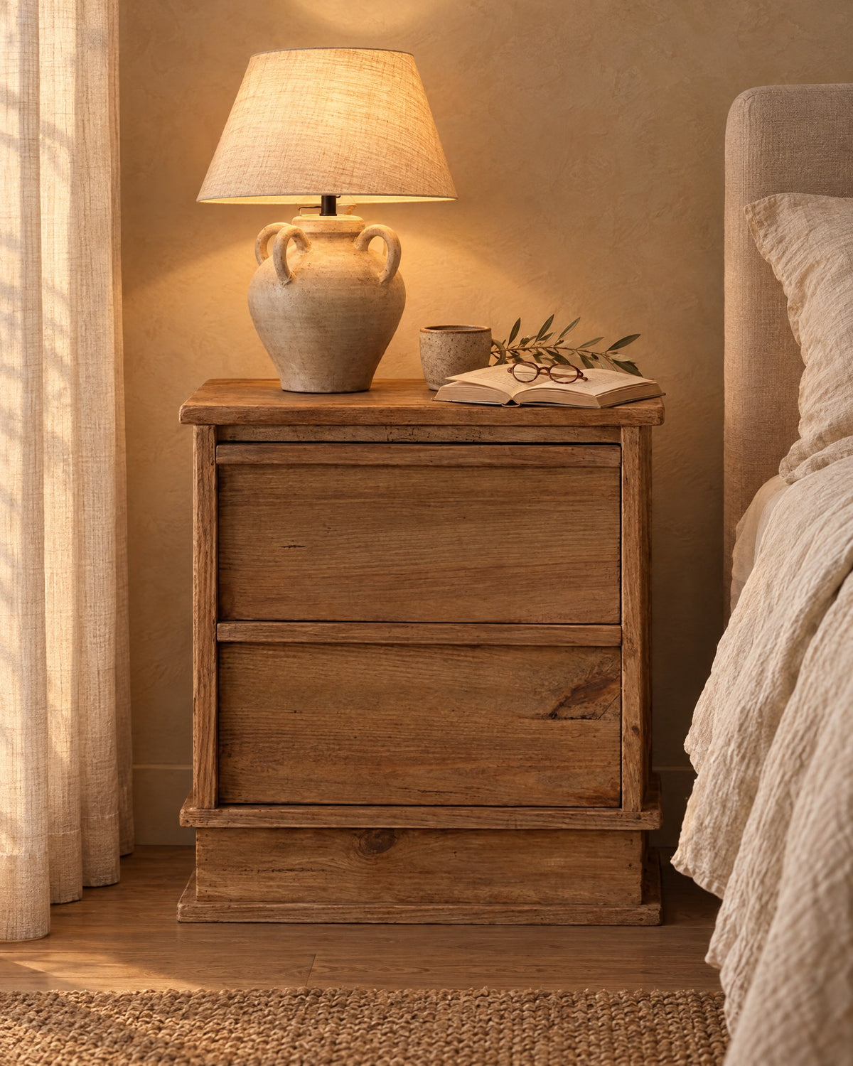 Rustic wood bedside table on a plinth base with a lamp switched on and an open book in the evening