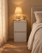 Slim cream bedside table with two drawers, a lamp switched on and an open book in the evening