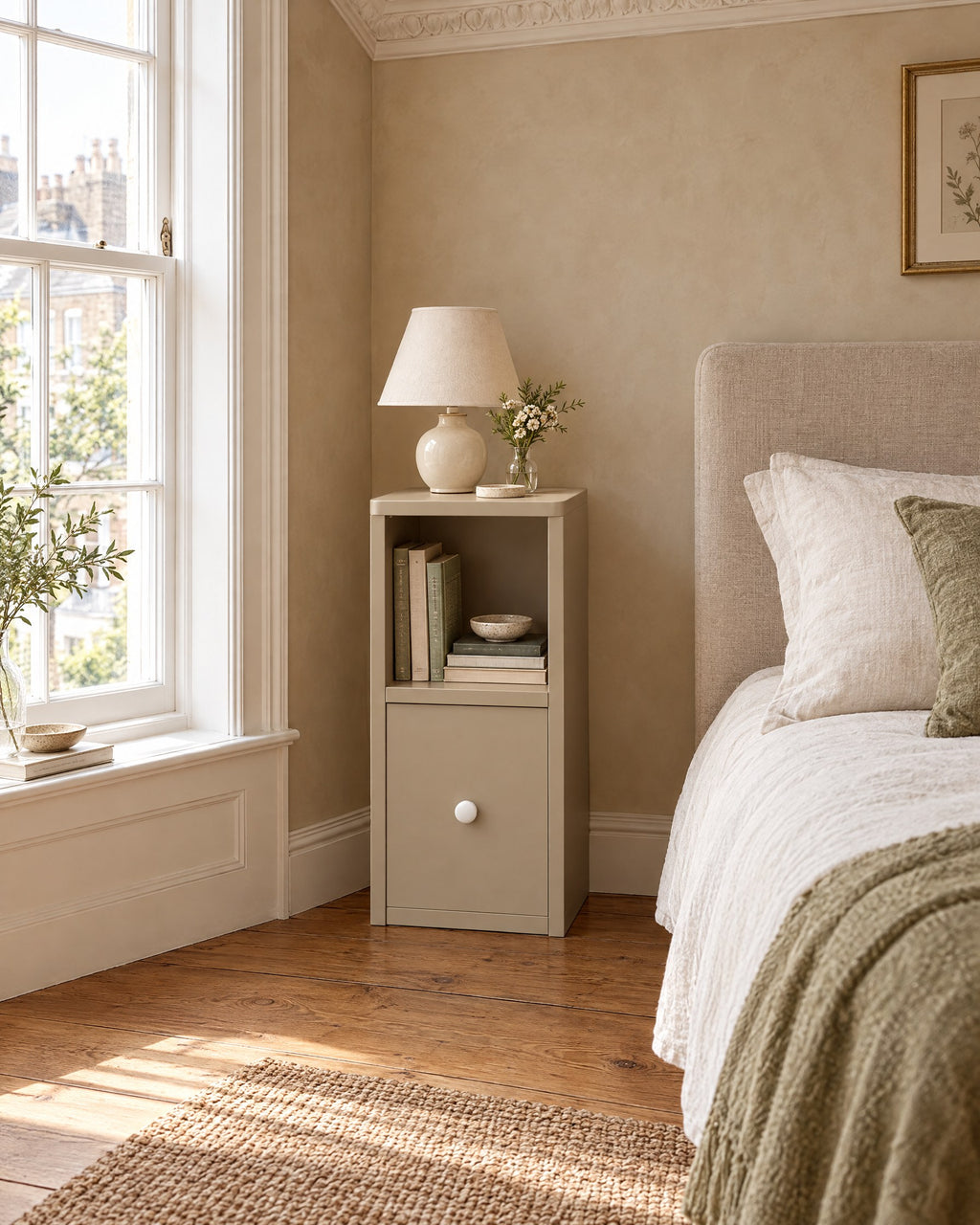 Slim greige bedside table with open shelf beside a linen bed in a bright British bedroom with a sash window