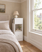 Slim 25 cm white bedside table beside a linen bed in a British bedroom with a sash window