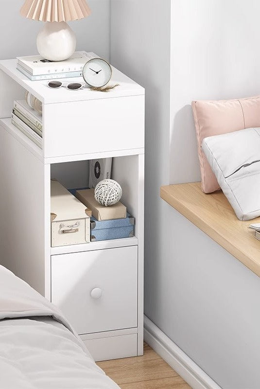 Slim white bedside table with side niche, open shelf and drawer beside a bed