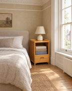 Small bamboo bedside table beside a linen bed in a bright British bedroom with a sash window