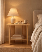 Small oak-effect bedside table with an open shelf, a lamp switched on and an open book in the evening