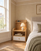 Small oak-effect bedside table with hutch shelf beside a linen bed in a bright British bedroom with a sash window