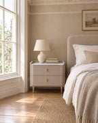 Taupe bedside table with gold legs beside a linen bed in a bright British bedroom with a sash window