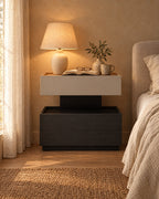 Two-tier greige and black bedside table with a lamp switched on and an open book in the evening