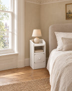 Ultra-narrow white bedside table beside a linen bed in a British bedroom with a sash window