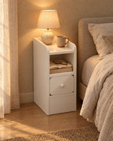 Ultra-narrow white bedside table with a lamp switched on and a book in the open shelf in the evening