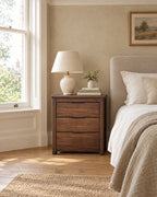 Walnut bedside table with three drawers and carved finger pulls beside a linen bed in a British bedroom with a sash window