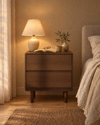 Walnut bedside table with flush drawers and a lamp switched on in the evening