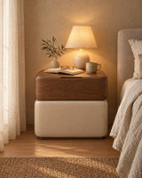 Walnut and cream upholstered bedside table with a lamp switched on and an open book in the evening