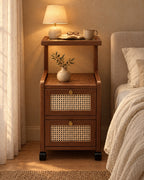 Walnut bedside table on wheels with cane drawers, a lamp switched on and an open book in the evening