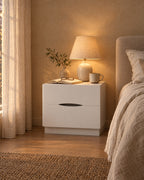 White bedside table with a curved finger pull, lamp switched on and an open book in the evening