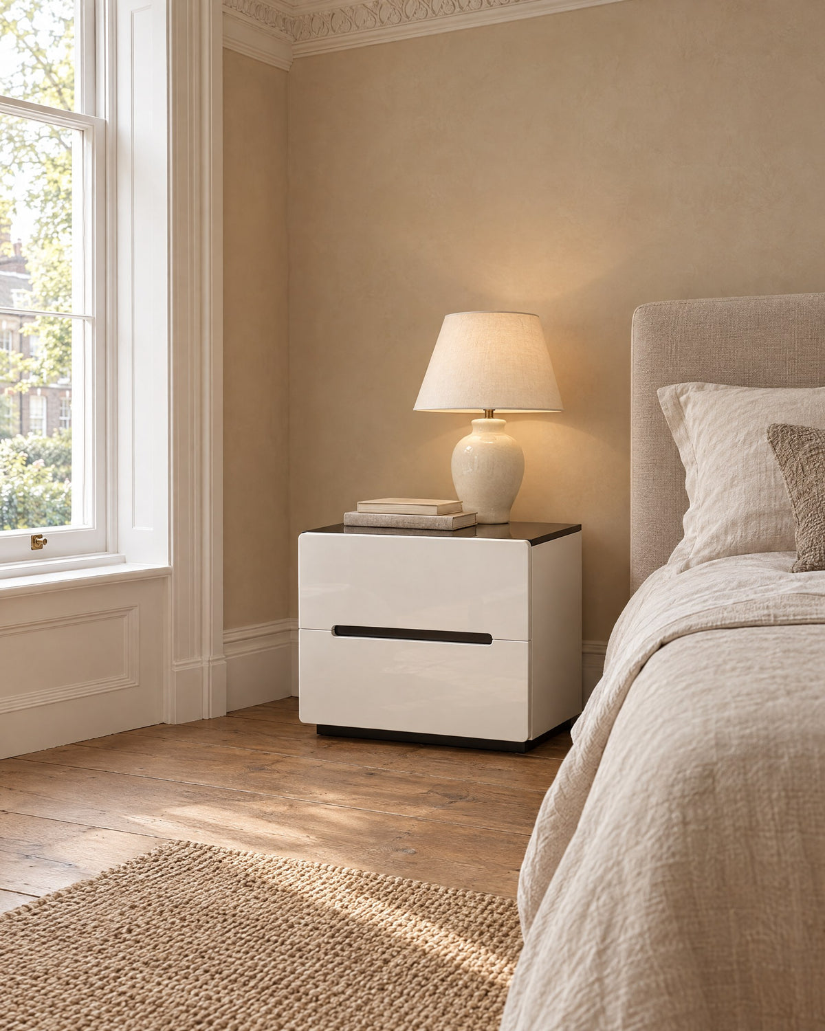 White gloss bedside table with a black top beside a linen bed in a British bedroom with a sash window