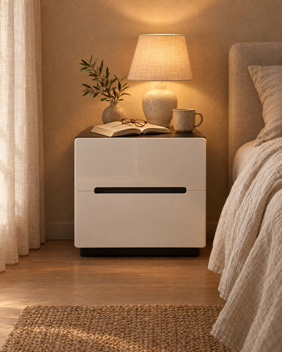 White gloss bedside table with a black top, lamp switched on and an open book in the evening