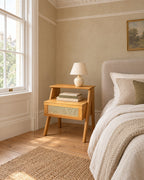 Wooden bedside table with a rattan drawer beside a linen bed in a British bedroom with a sash window